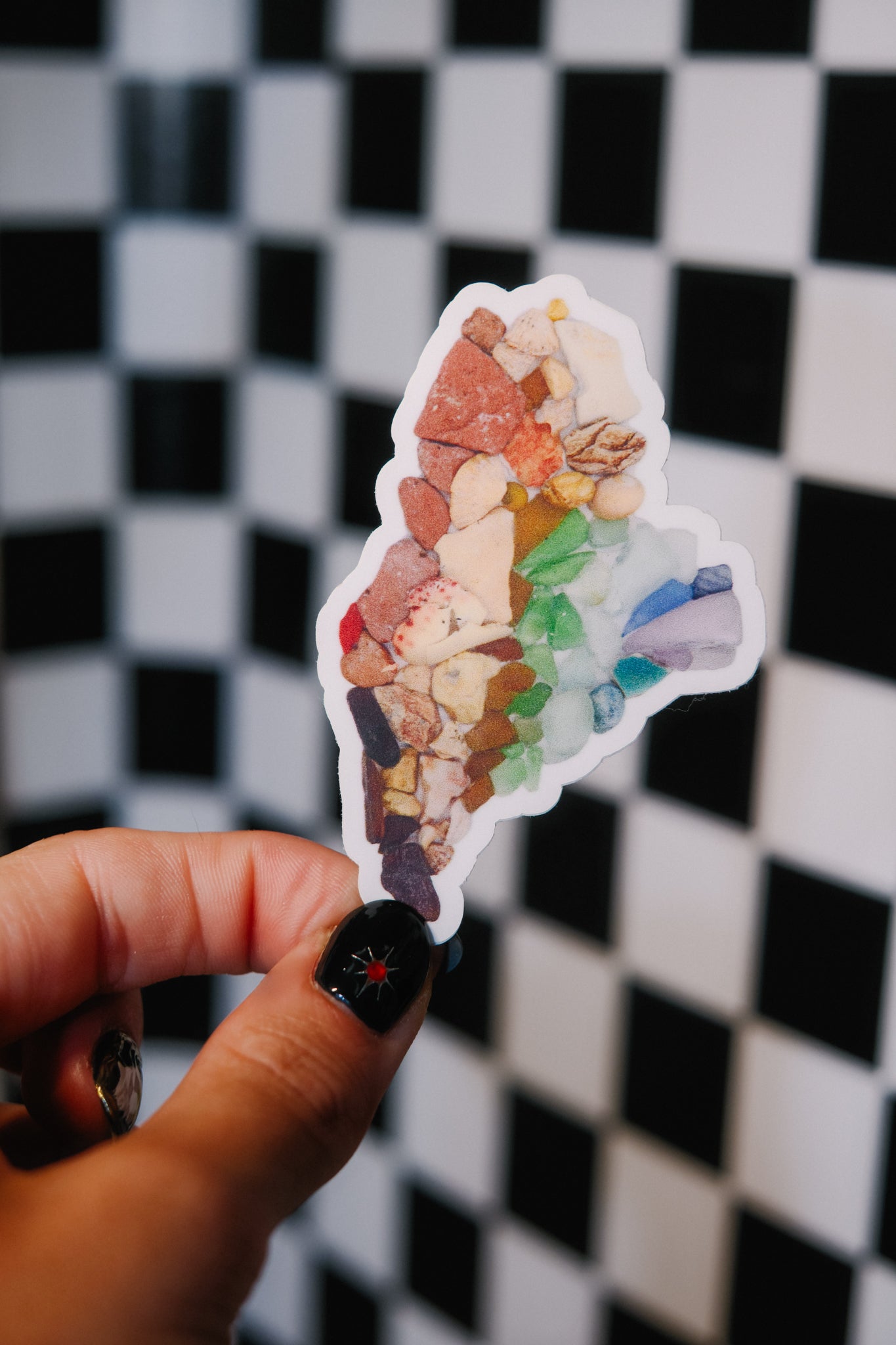 Colorful gemstone sticker in the shape of the state of maine. Held by a hand against a black and white checkered background