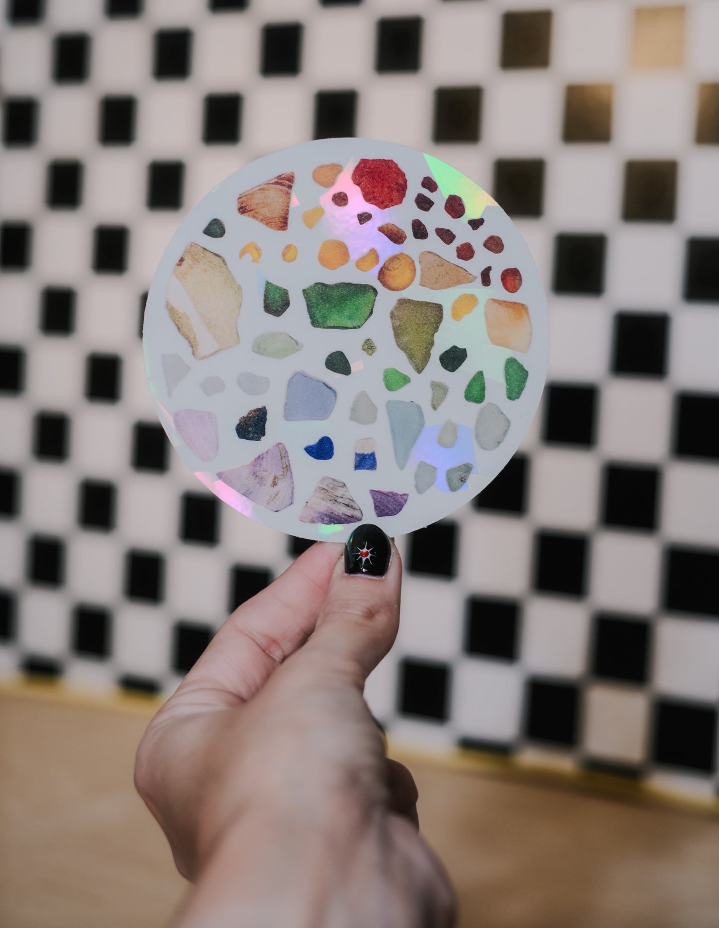 Hand holding a decal suncatcher comprised of rainbow sea glass pieces, with a black and white checkered background.