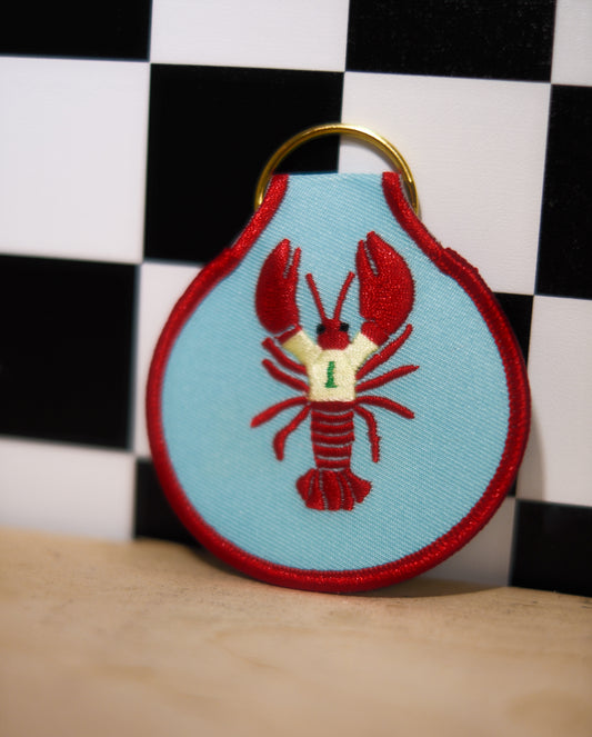 Keychain with red lobster design on a light blue background against a checkered black and white wall.