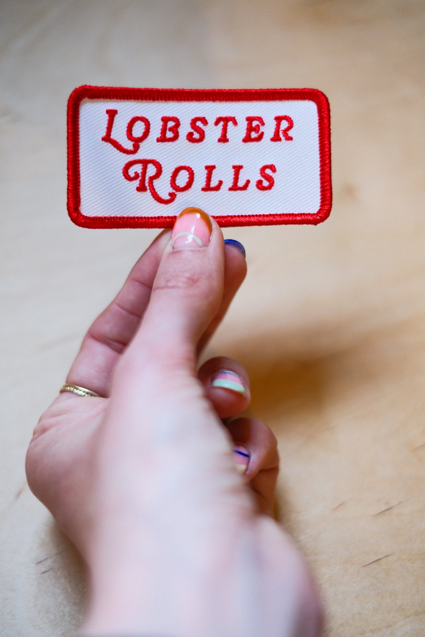 Hand holding a patch with 'LOBSTER ROLLS' in red text on a wooden surface.