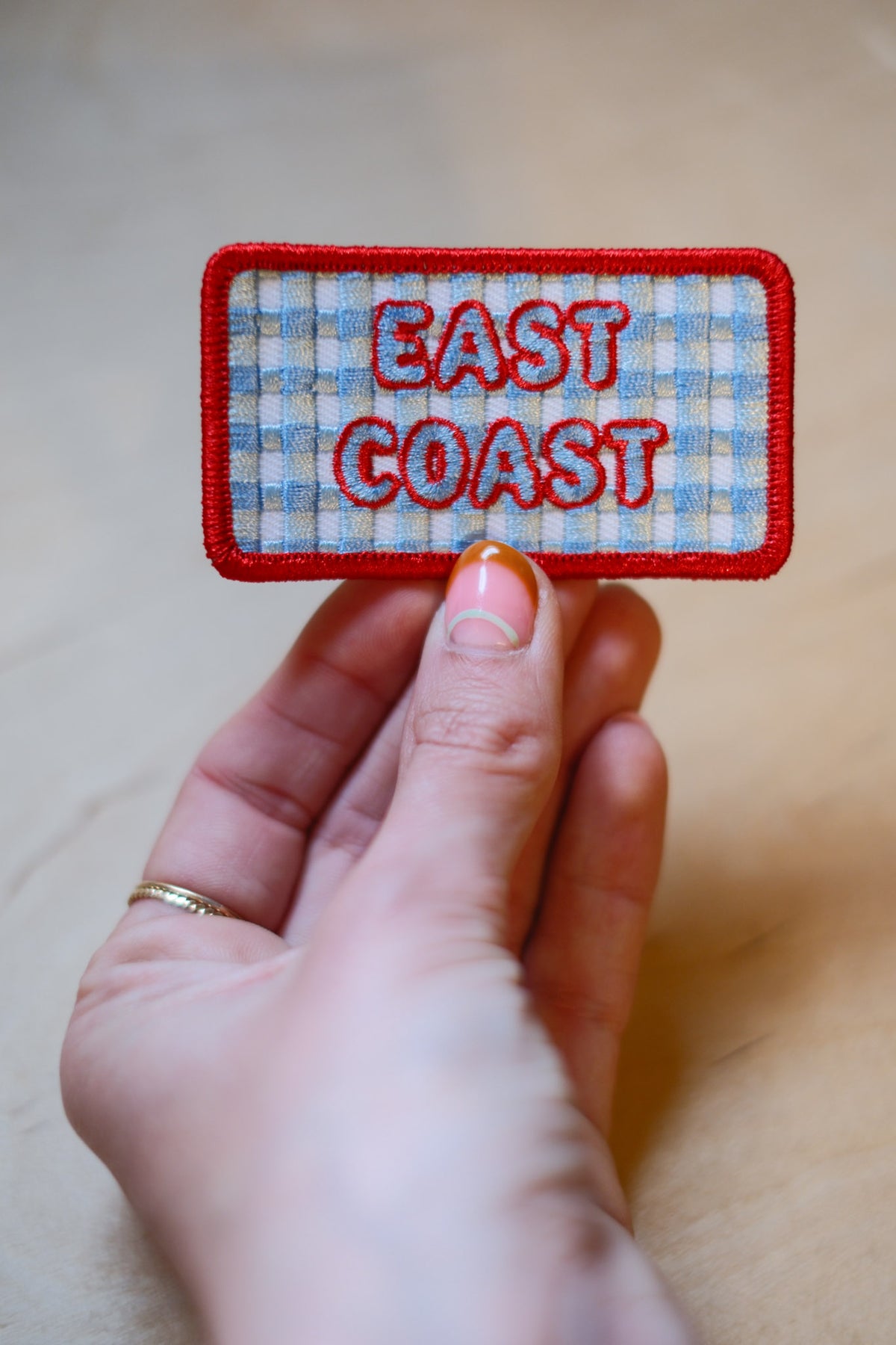 Hand holding a patch with that says 'East Coast' featuring blue and white checkered background and red embroidered border.