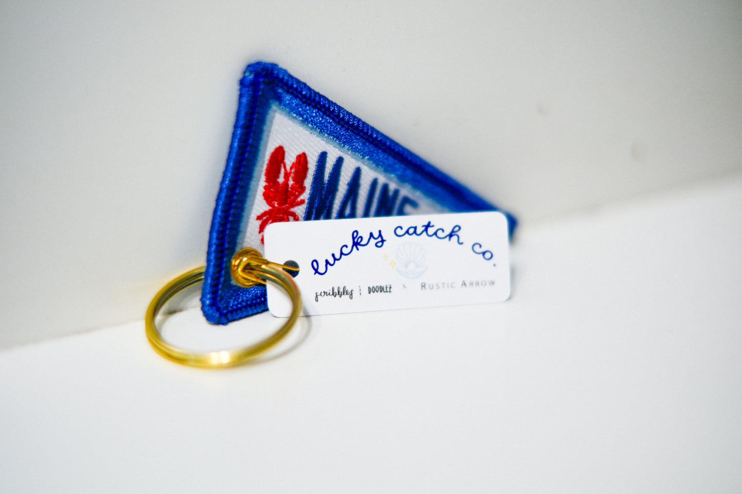 A triangular embroidered patch keychain with a blue border and white center, featuring a red lobster and blue lettering"MAINE" attached is white tag that reads 'lucky catch co.' and a gold split ring, photographed on a white surface.