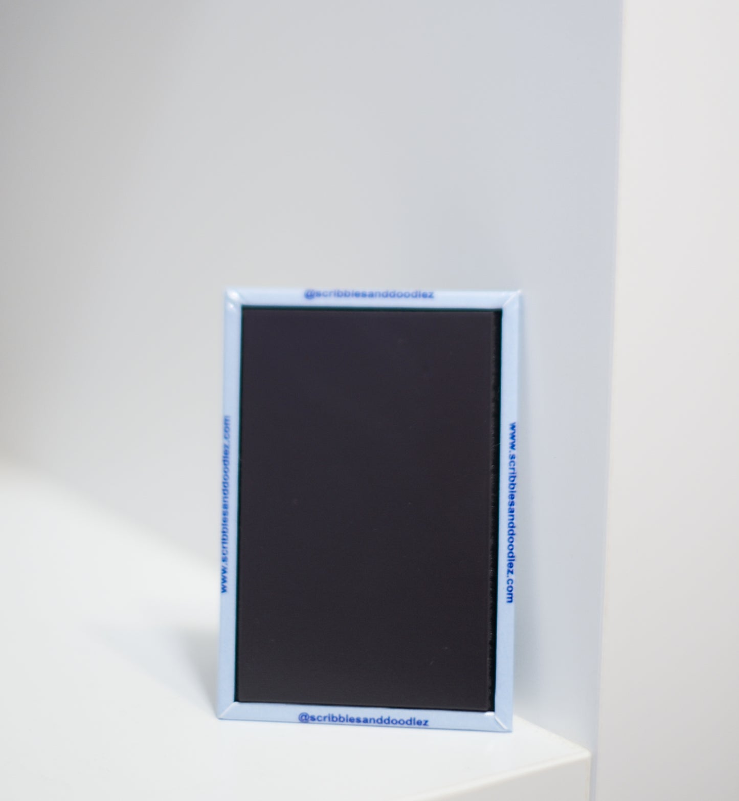 A small rectangular magnet with a light blue border stands upright on a white surface against a white background. Blue text is printed along the border, including 'www.scribblesanddoodlez.com' and '@scribblesanddoodlez'