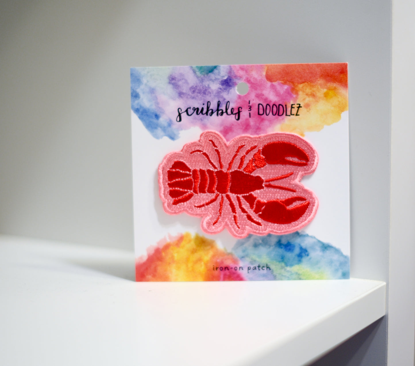 A red lobster-shaped iron-on patch with pink bordering, displayed on a white backing card with a colorful watercolor design. The card shows the brands 'scribbles & doodlez" and 'iron-on patch' and is photographed on a white shelf against a white background.