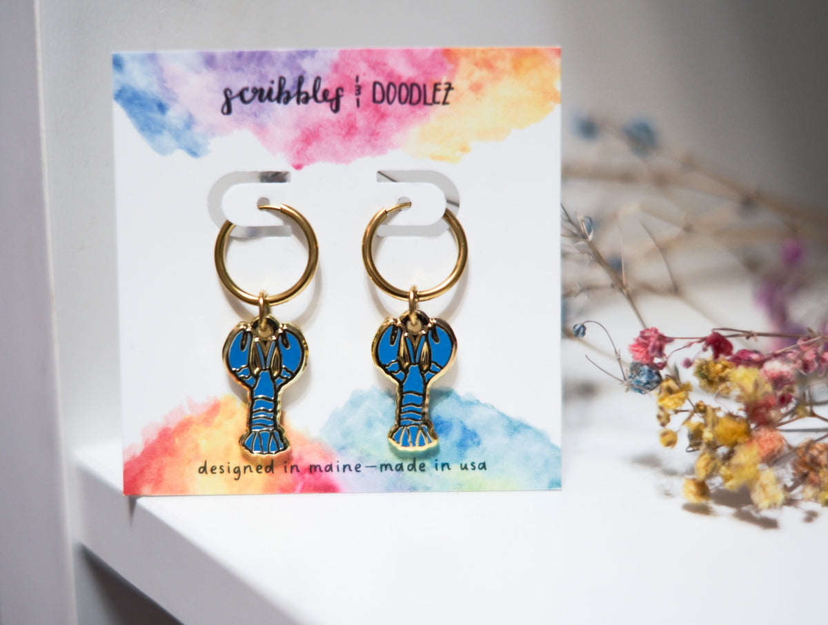 A pair of gold-tone lobster-shaped hoop earrings with blue enamel detailing, displayed on a white card with a colorful watercolor background. The card is branded 'scribbles + doodlez' and the earrings are photographed on a white surface with blurred dried flowers in the background.