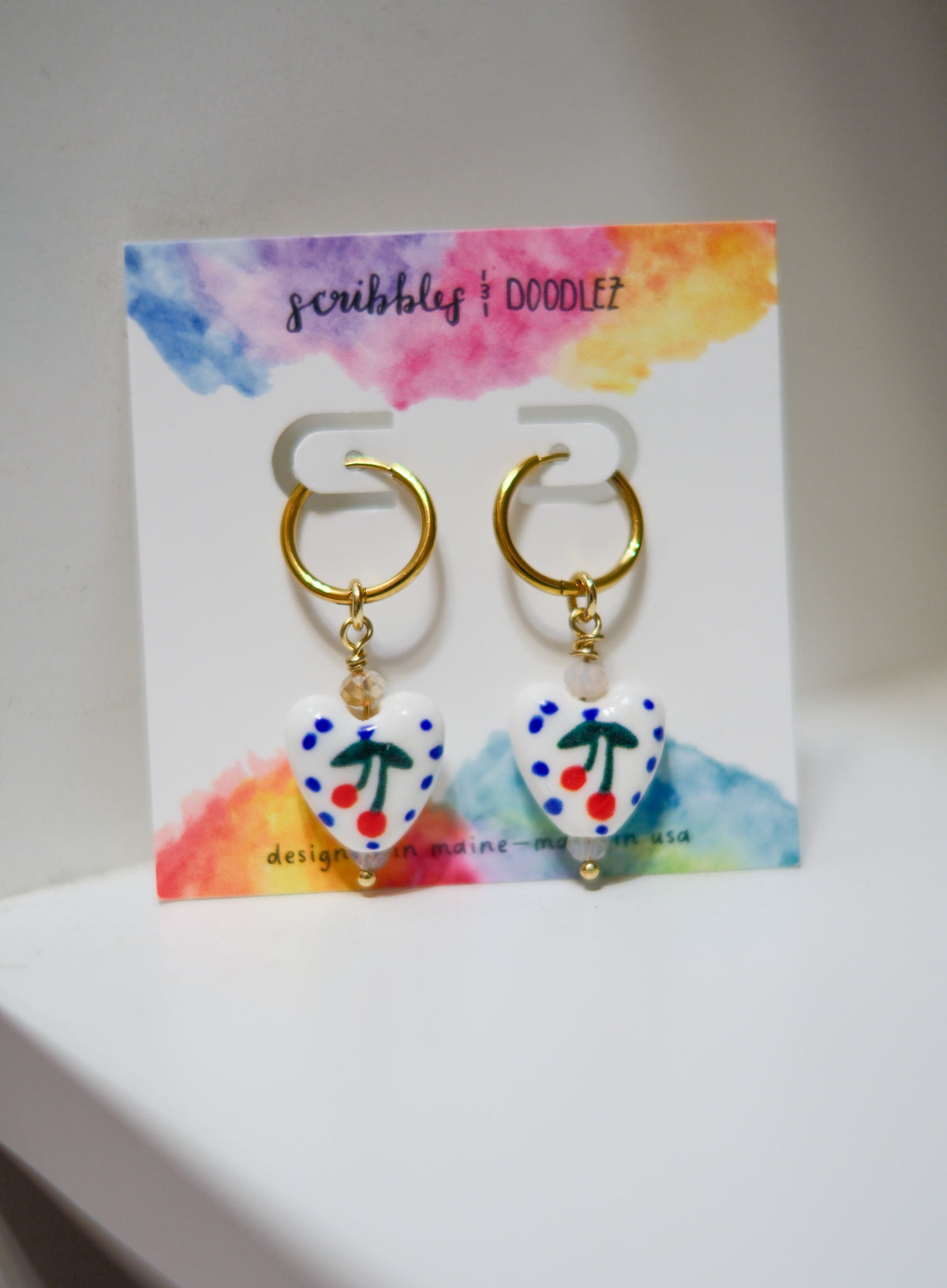 A pair of gold hoop earrings with dangling white heart beads decorated with blue polka dots and red cherries, displayed on a colorful watercolor card labeled 'scribbles & doodlez'