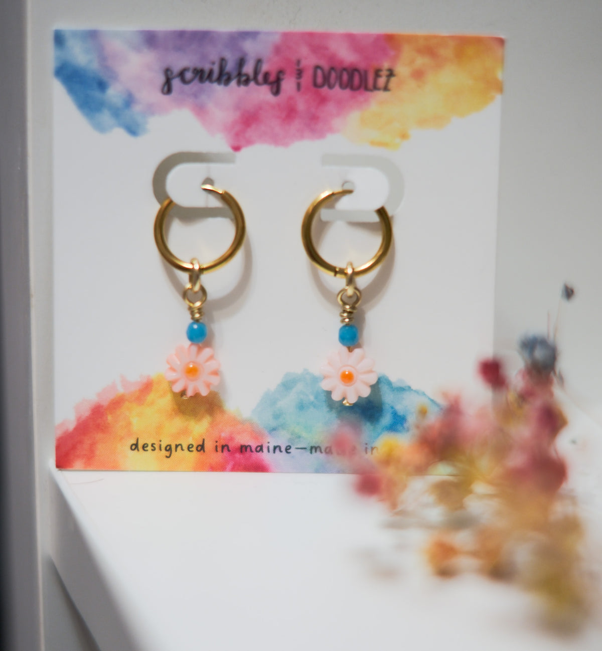 A pair of gold huggie hoop earrings with small blue beads and pink daisy shell beads, displayed on a colorful card that reads 'scribbles & doodlez.