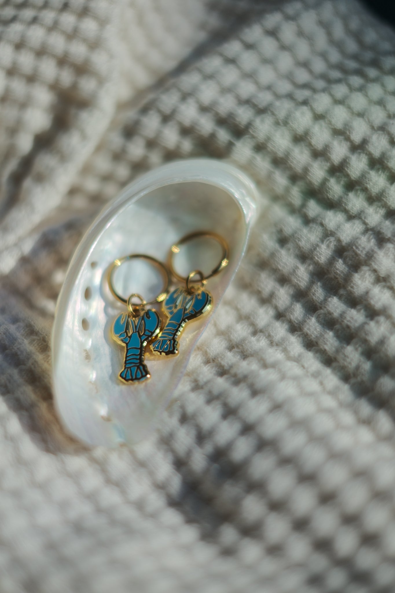 A pair of lobster-shaped drop earrings with gold-tone metal and turquoise blue enamel detailing, resting inside a white shell on a textured beige fabric background.