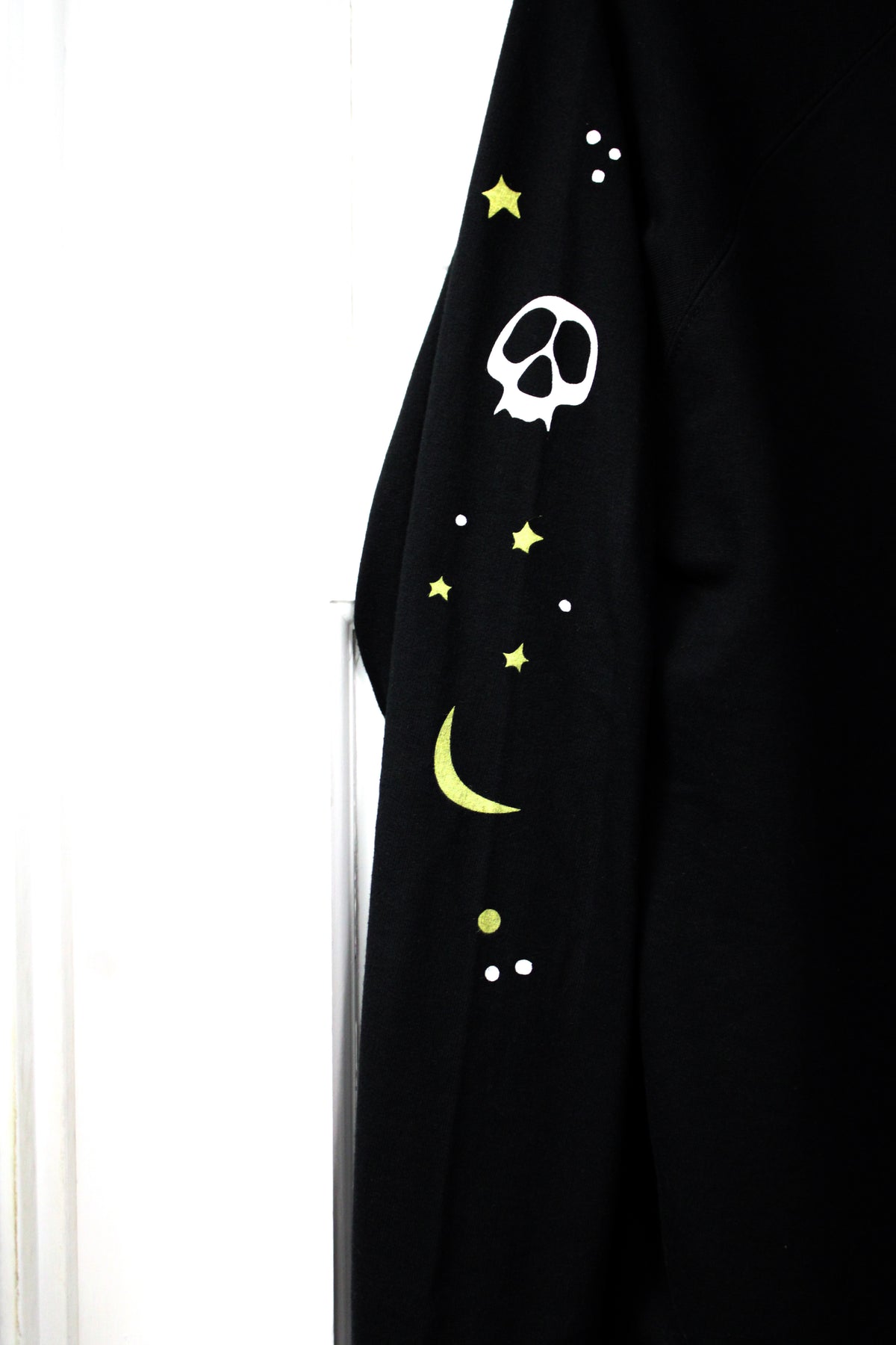detail shot of sleeve of a black crew neck sweatshirt, featuring yellow stars and moons, and white skeleton faces.