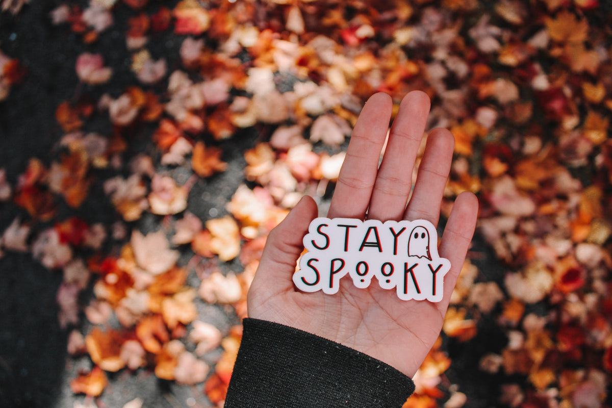 A hand holding a die-cut sticker that says 'STAY SPOOKY' with a small ghost icon, set against a background of scattered autumn leaves.