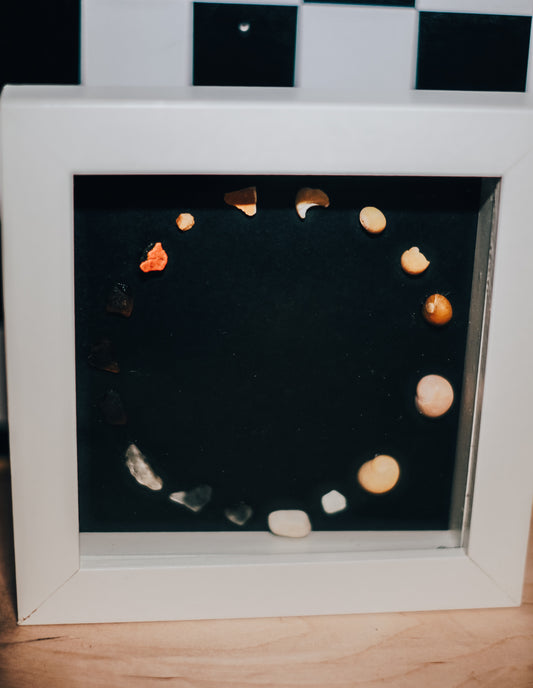 Decorative shadow box with pebbles arranged in a circular pattern on a dark background.