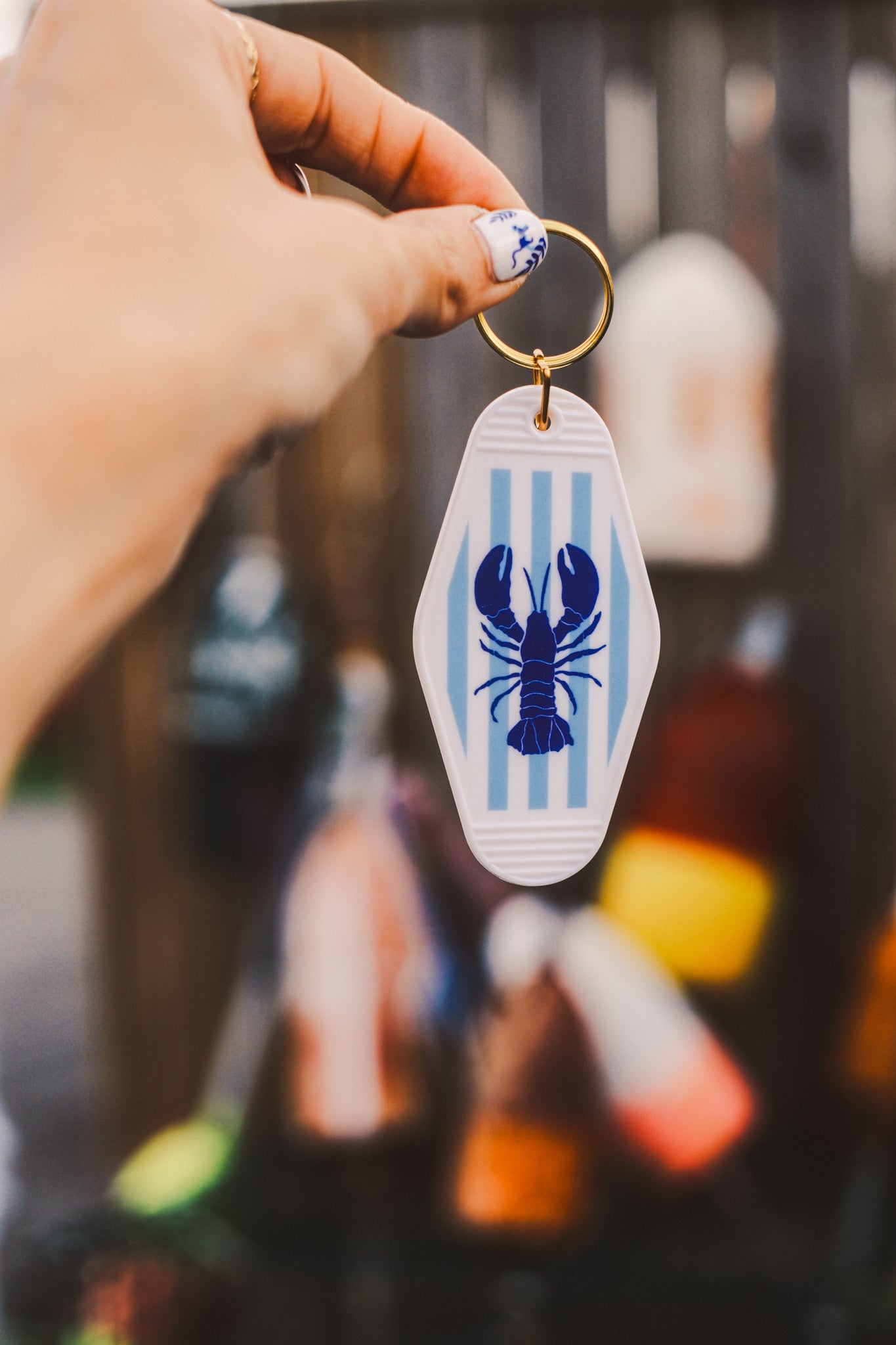 Keychain with blue lobster illustration, background of keychain blue and white stripes. Held by a hand against a blurred background.