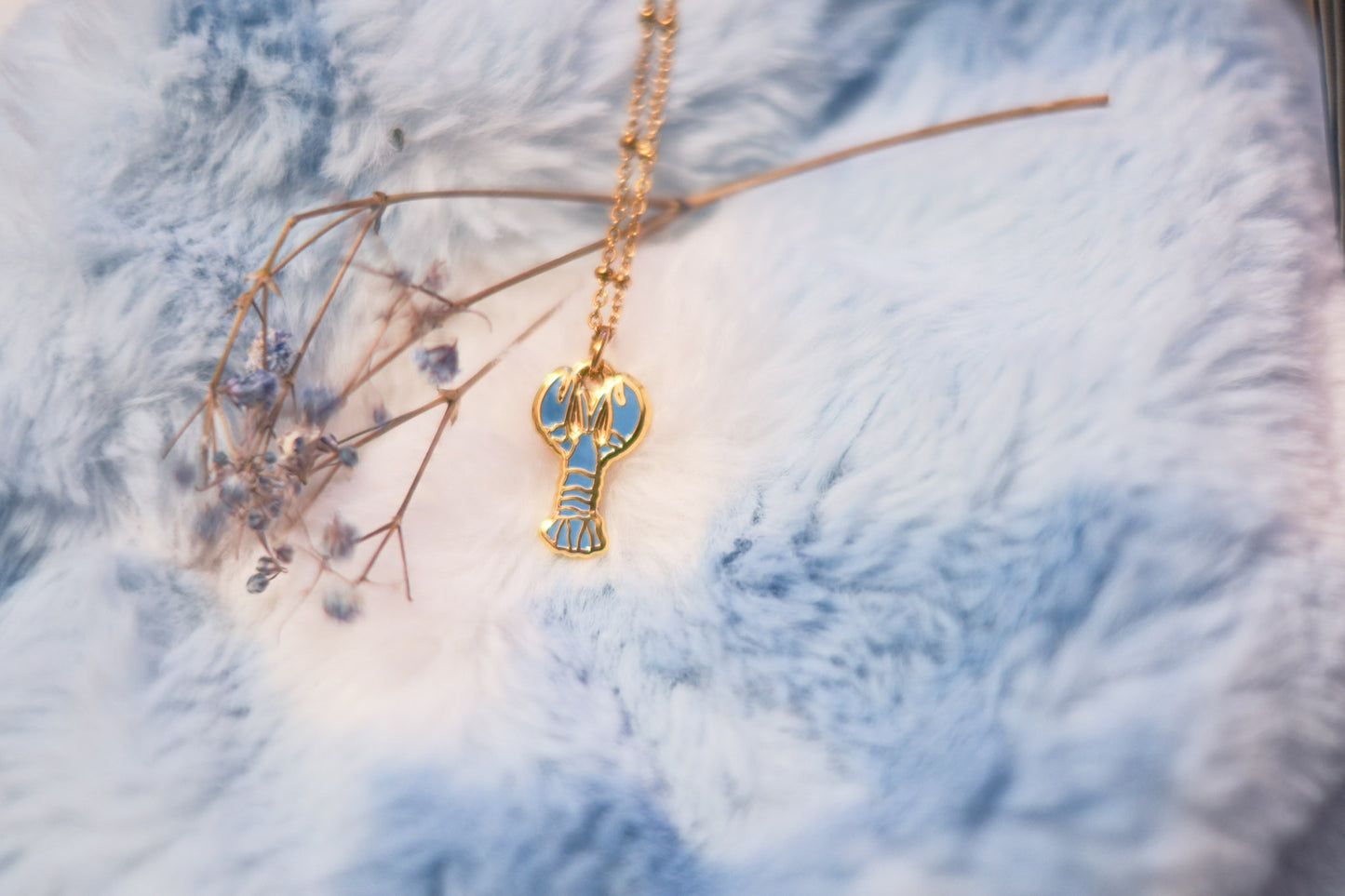 Gold necklace with a blue lobster pendant on a soft blue background.