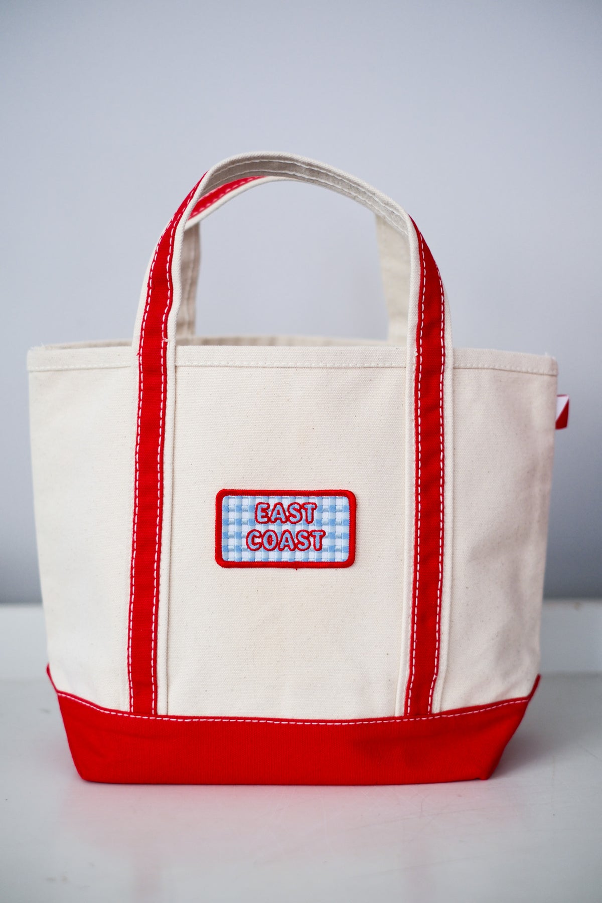 A canvas tote bag with cream-colored fabric, red trim, and a red bottom panel, featuring a front patch that reads 'EAST COAST' on a blue-and-white gingham background.
