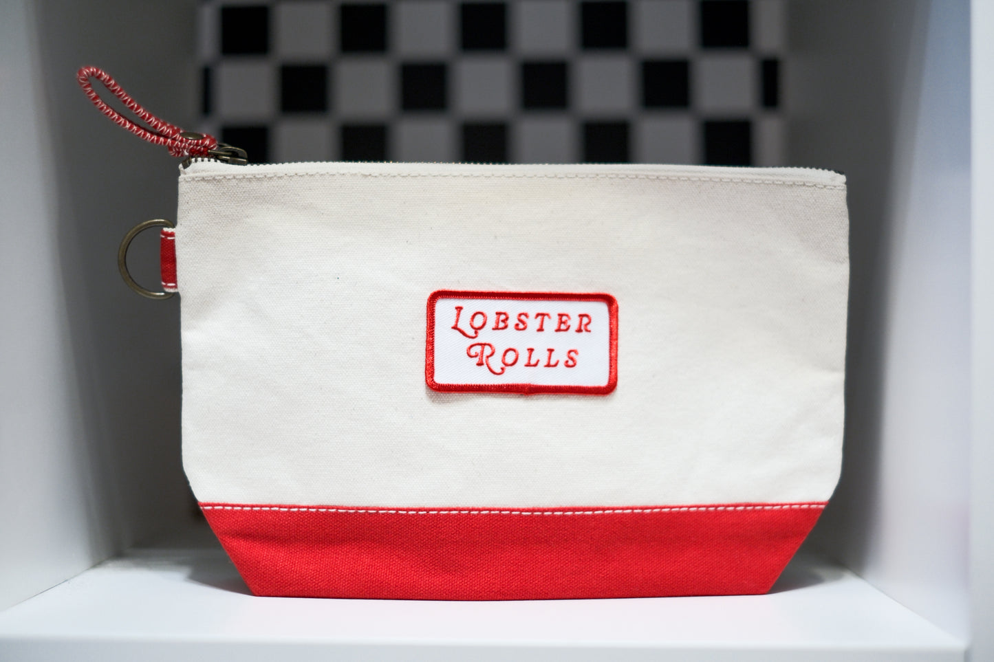 A canvas zippered pouch with an off-white top and a red bottom panel, featuring a red rectangular patch with the words 'LOBSTER ROLLS' in red script. A red zipper pull and metal ring are attached on the left side.