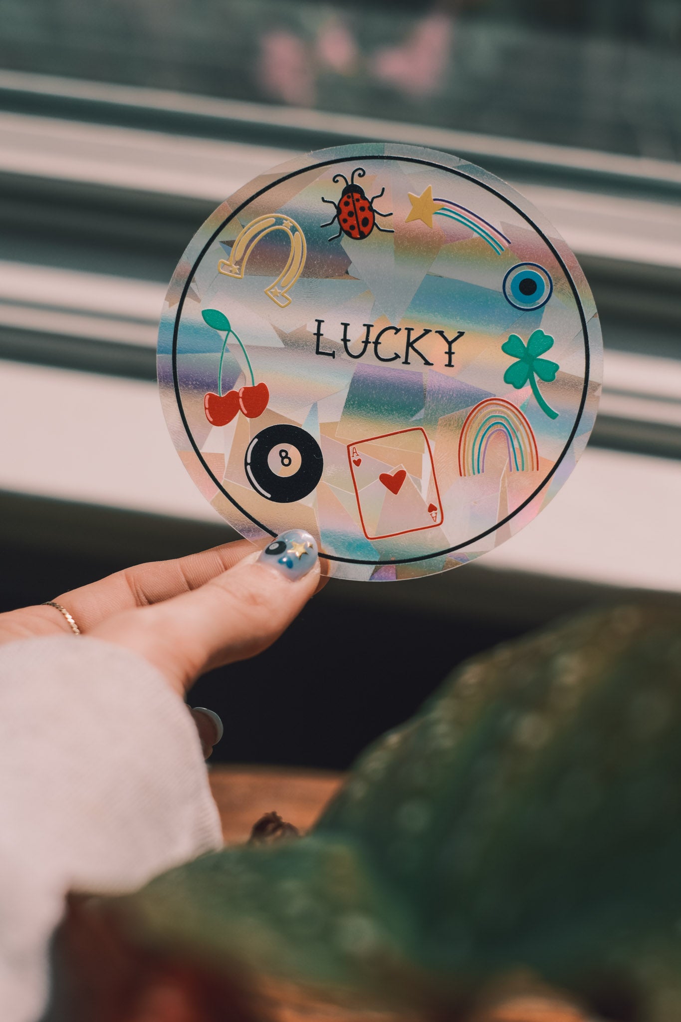 Hand holding a round sun catcher decal with various good luck symbols and the word 'LUCKY' on a blurred background.