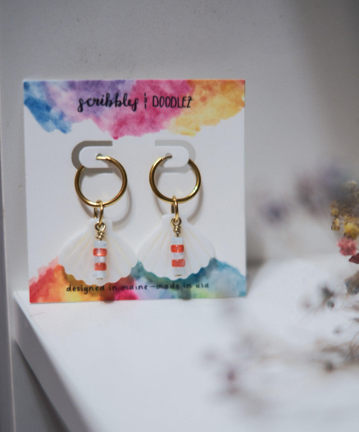 A pair of gold hoop earrings with white scallop charms and red and blue featuring red and blue beads displayed on a branded 'scribbles & doodlez' backing card.