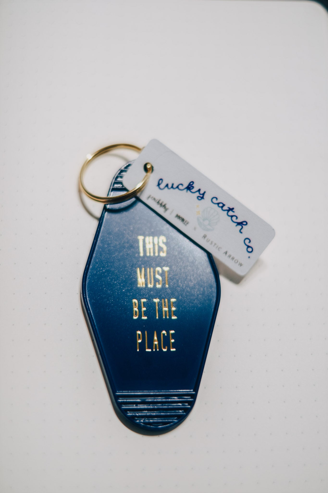 Blue keychain with 'This must be the place' text and a tag on a light background