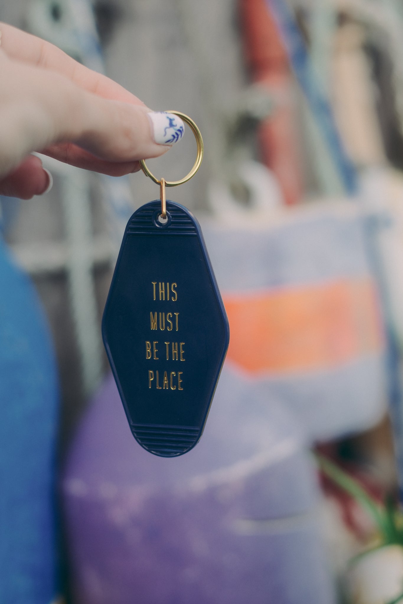 Navy blue keychain with 'THIS MUST BE THE PLACE' text held by a hand against a blurred background.