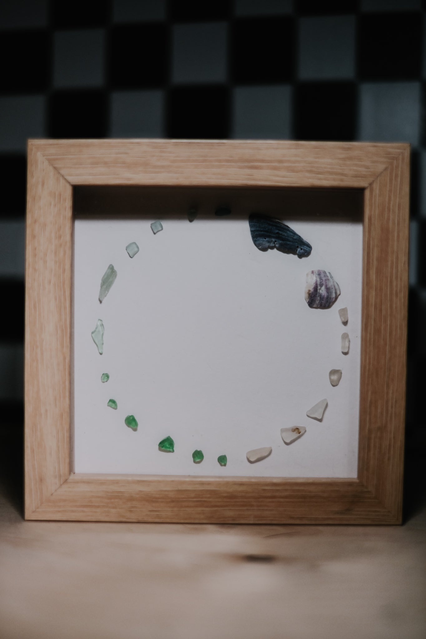 Framed artwork with small shells and sea glass arranged in a circular pattern on a white background.