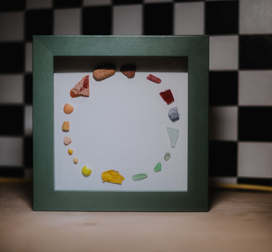 Framed artwork with colorful shells and glass arranged in a circular pattern on a white background.