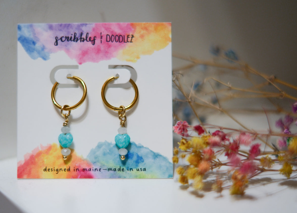 Gold huggie hoop earrings with turquoise heart beads and aquamarine crystal beads, displayed on a colorful watercolor backing card that reads 'scribbles & doodlez'