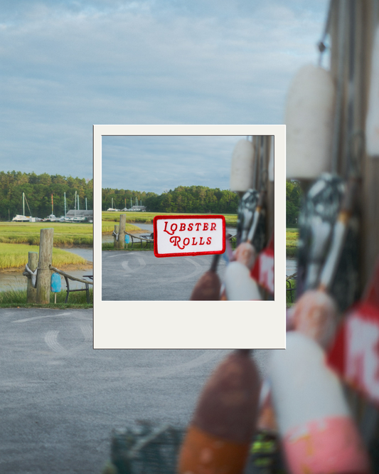 A Polaroid-style photo centered in the frame, showing a small white iron on patch with red text that reads 'LOBSTER ROLLS' in the background is a marsh, with buoys, boats, and water.