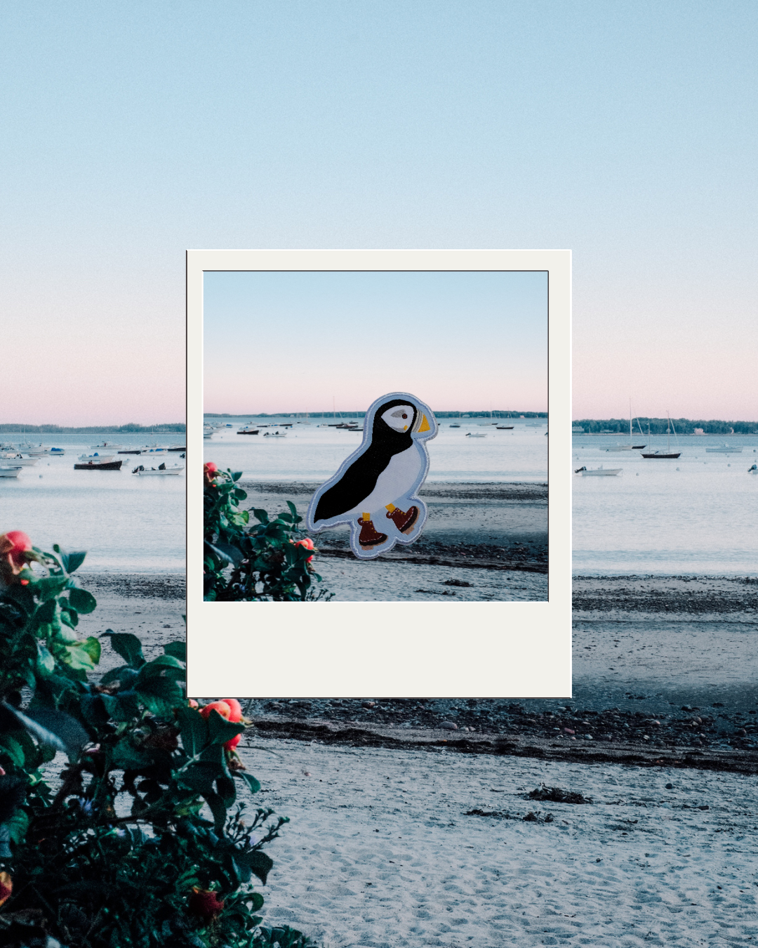 A die-cut iron on patch of a puffin standing on a beach with brown boots presented inside a white polaroid-style frame against a scenic beach background at sunset.