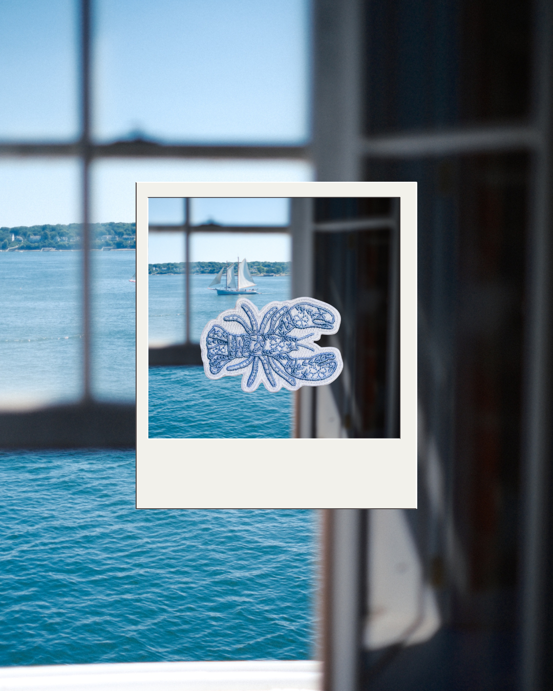 A iron on patch featuring a white lobster design with blue floral detailing, placed on a Polaroid-style photo frame that is set against a real-world window view of blue water and a sailboat.