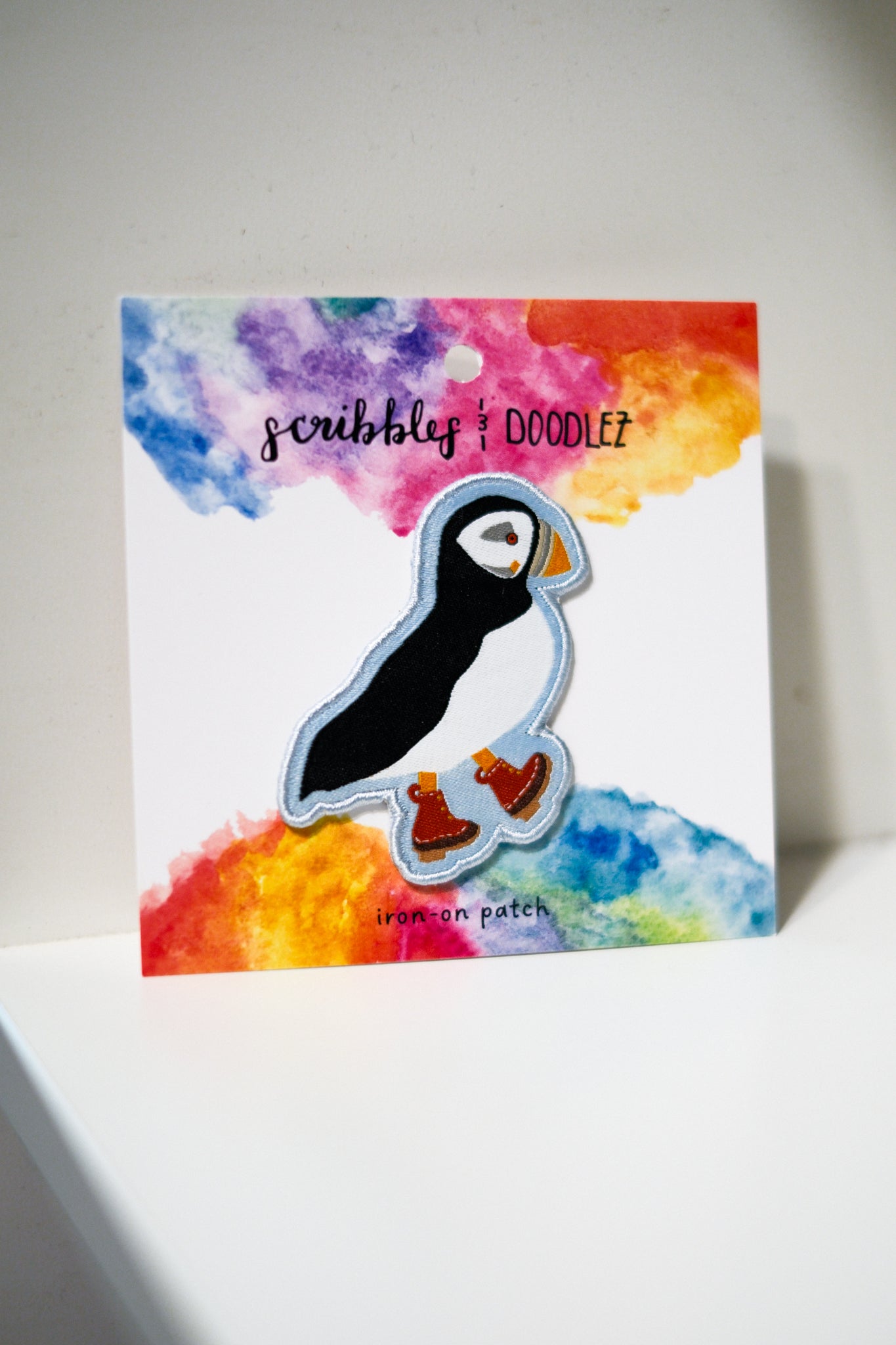 A puffin-shaped iron-on patch with a black and white body, orange beak, and orange feet, displayed on a colorful watercolor backing card.