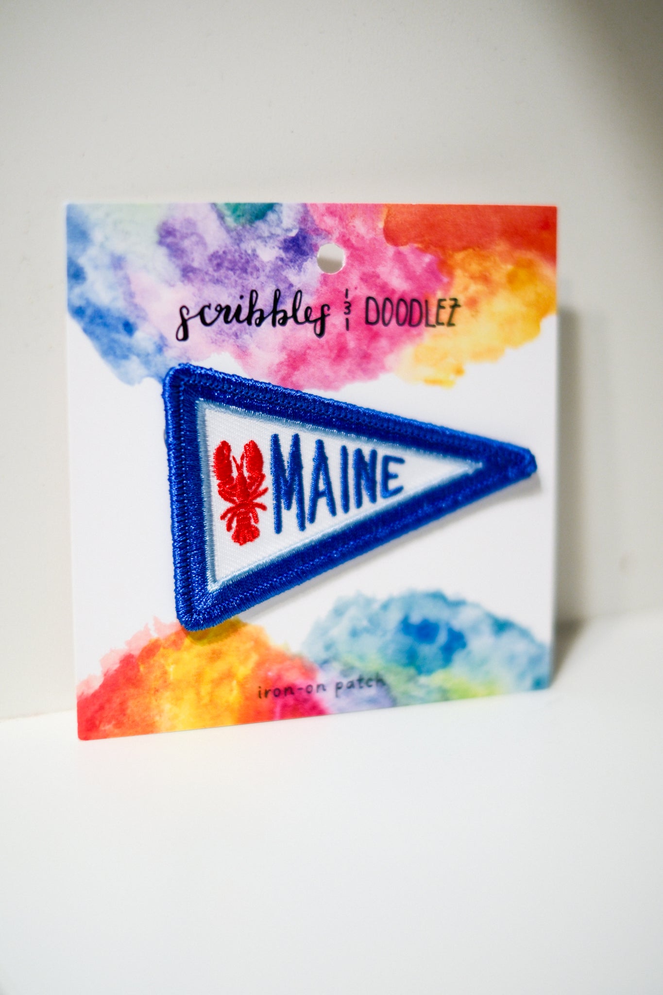A triangular embroidered patch featuring the word 'MAINE' in blue with a red lobster icon, mounted on a white backing card.