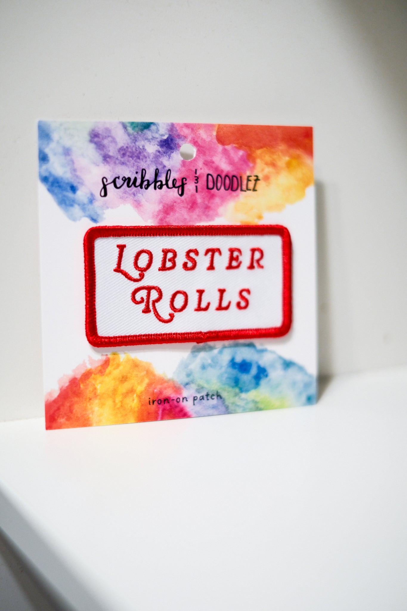 A square embroidered patch reading 'LOBSTER ROLLS' in red lettering inside a red border, mounted on a white card with colorful watercolor-style artwork and the text 'scribbles & DOODLEZ' and 'iron-on patch'.