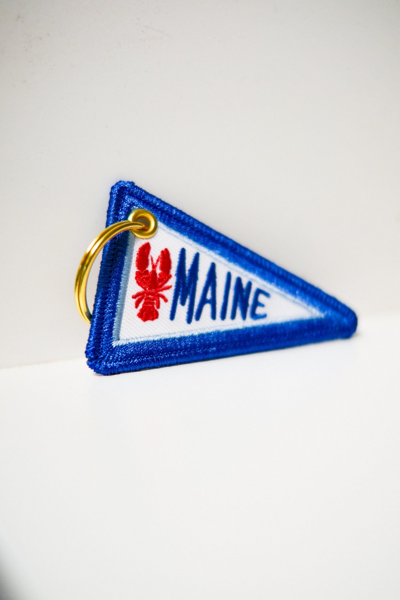 A triangular embroidered keychain featuring a blue border, a white center, a red lobster, and the word 'MAINE' in blue, attached to a gold metal key ring.