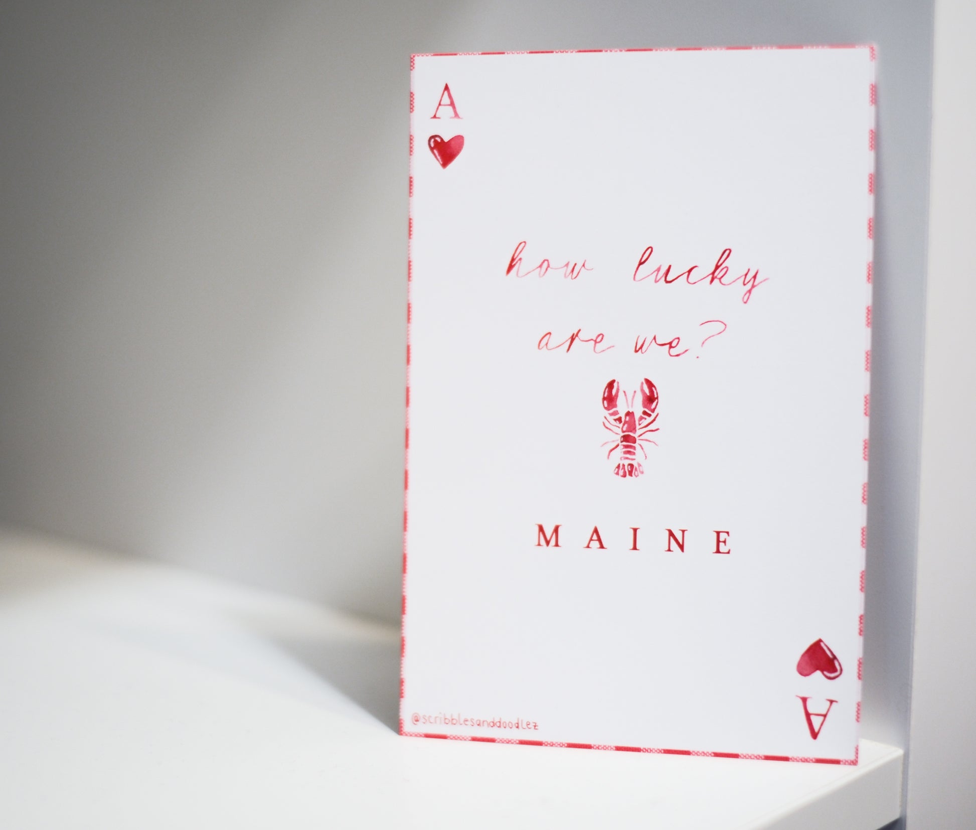 A white greeting card styled like an Ace of Hearts playing card, featuring red text that reads 'how lucky are we?' and 'MAINE' with a lobster illustration, standing on a white surface against a gray background.