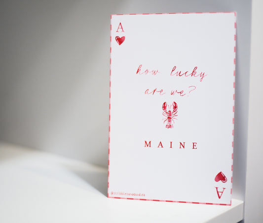 A white greeting card styled like an Ace of Hearts playing card, featuring red text that reads 'how lucky are we?' and 'MAINE' with a lobster illustration, standing on a white surface against a gray background.