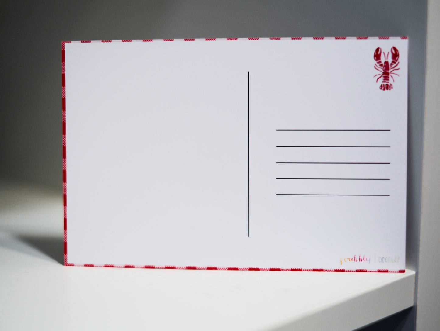 A white postcard with a red dashed border, a red lobster illustration in the top right, and writing lines on the right side, standing on a white surface against a gray background.
