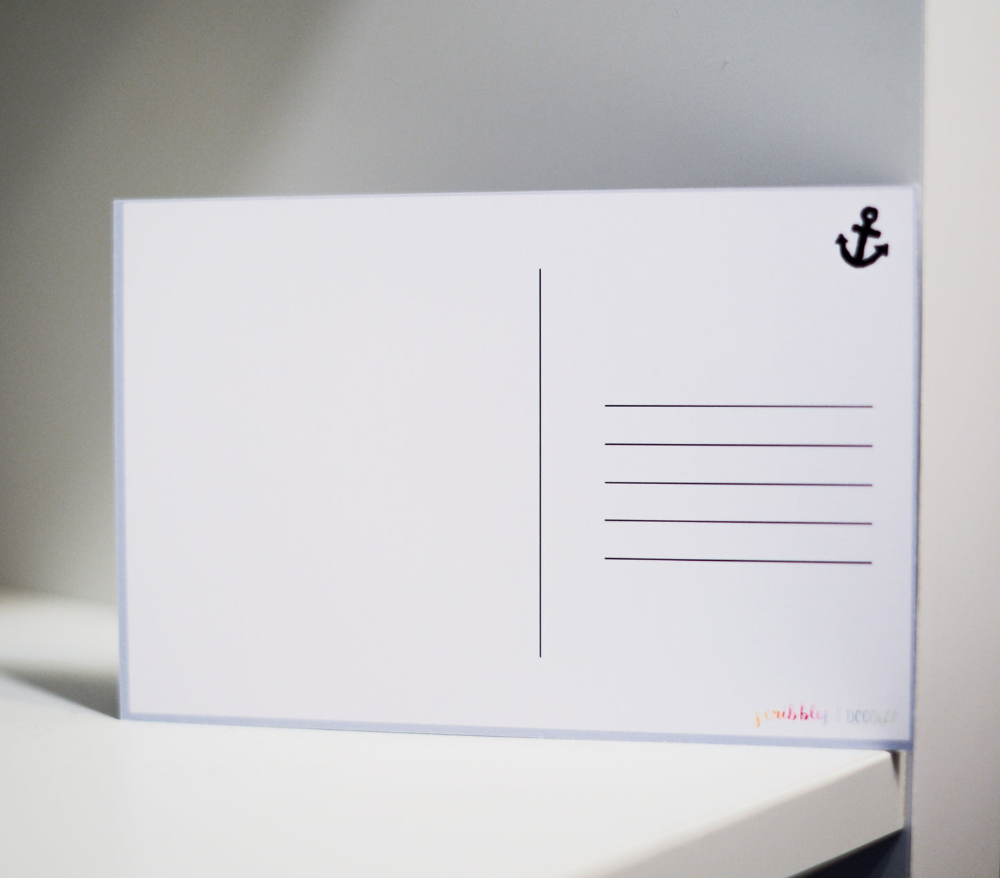 A white postcard with a black anchor icon in the top right corner and printed postcard lines on the right side, resting on a white surface against a gray background.