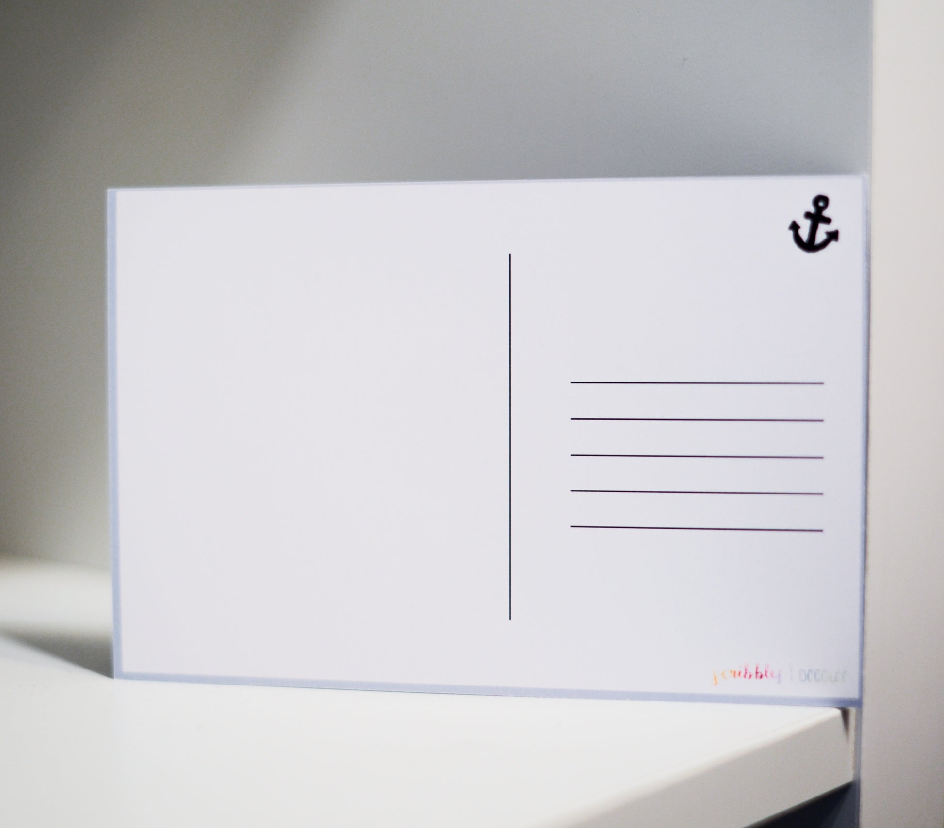 A white postcard with a black anchor icon in the top right corner and printed postcard lines on the right side, resting on a white surface against a gray background.