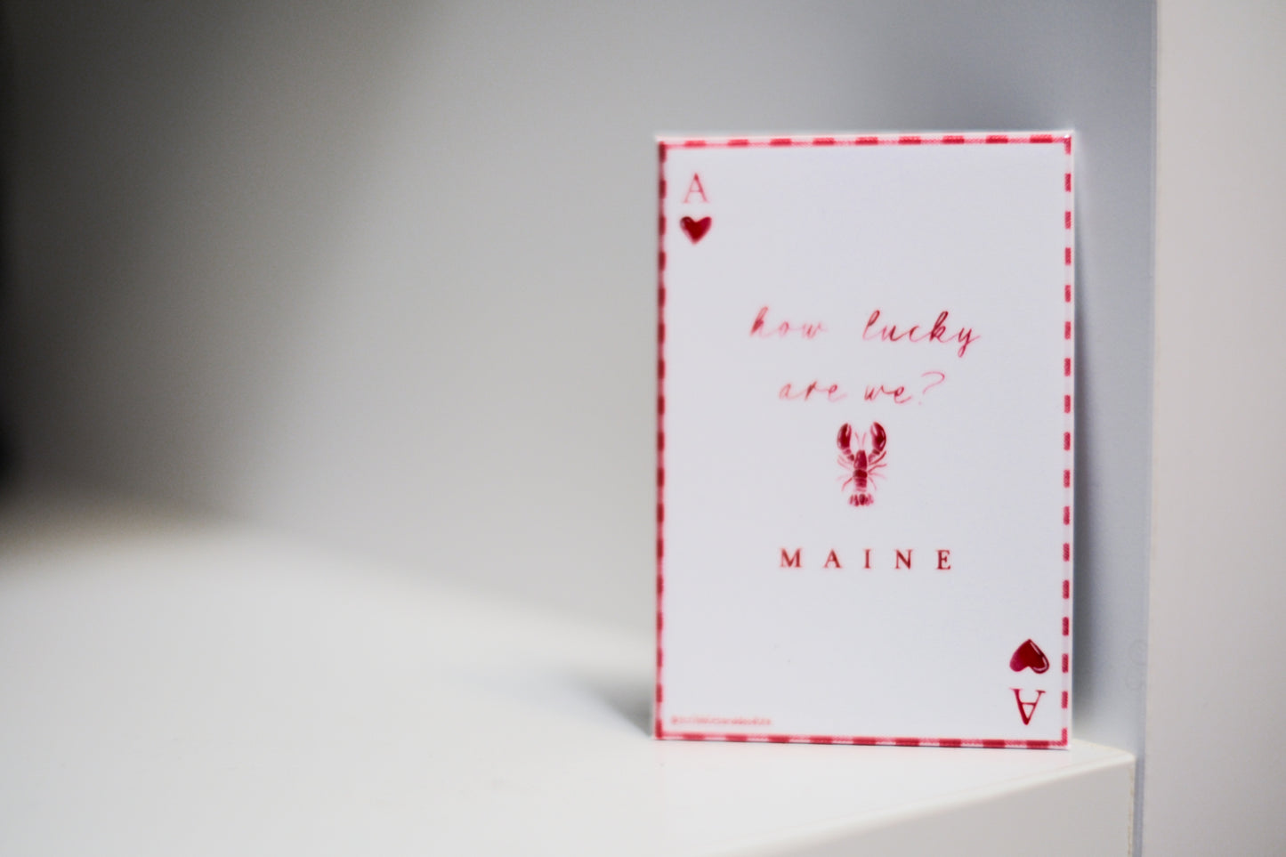 A white magnet with a red and white plaid border. The magnet is styled like an Ace of Hearts, red heart symbols in the corners, and red script text reading 'how lucky are we?' with 'MAINE' and a lobster graphic below.
