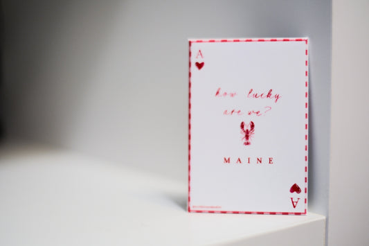 A white magnet with a red and white plaid border. The magnet is styled like an Ace of Hearts, red heart symbols in the corners, and red script text reading 'how lucky are we?' with 'MAINE' and a lobster graphic below.