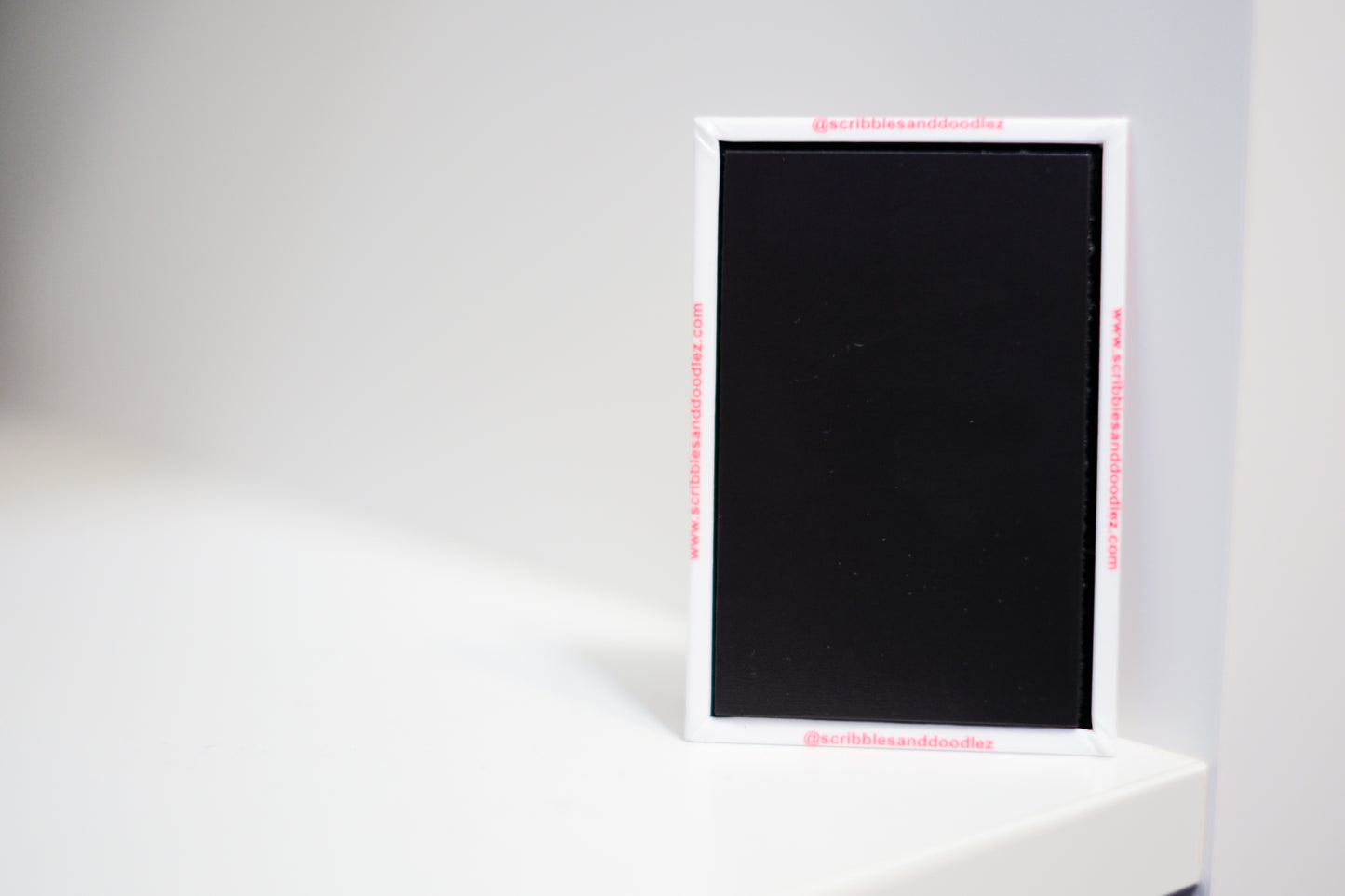 A square black magnet with a white border, standing on a white surface against a light gray background. The magnet has red text printed on the border, '@scribblesanddoodlez' 'www.scribblesanddoodlez.com'