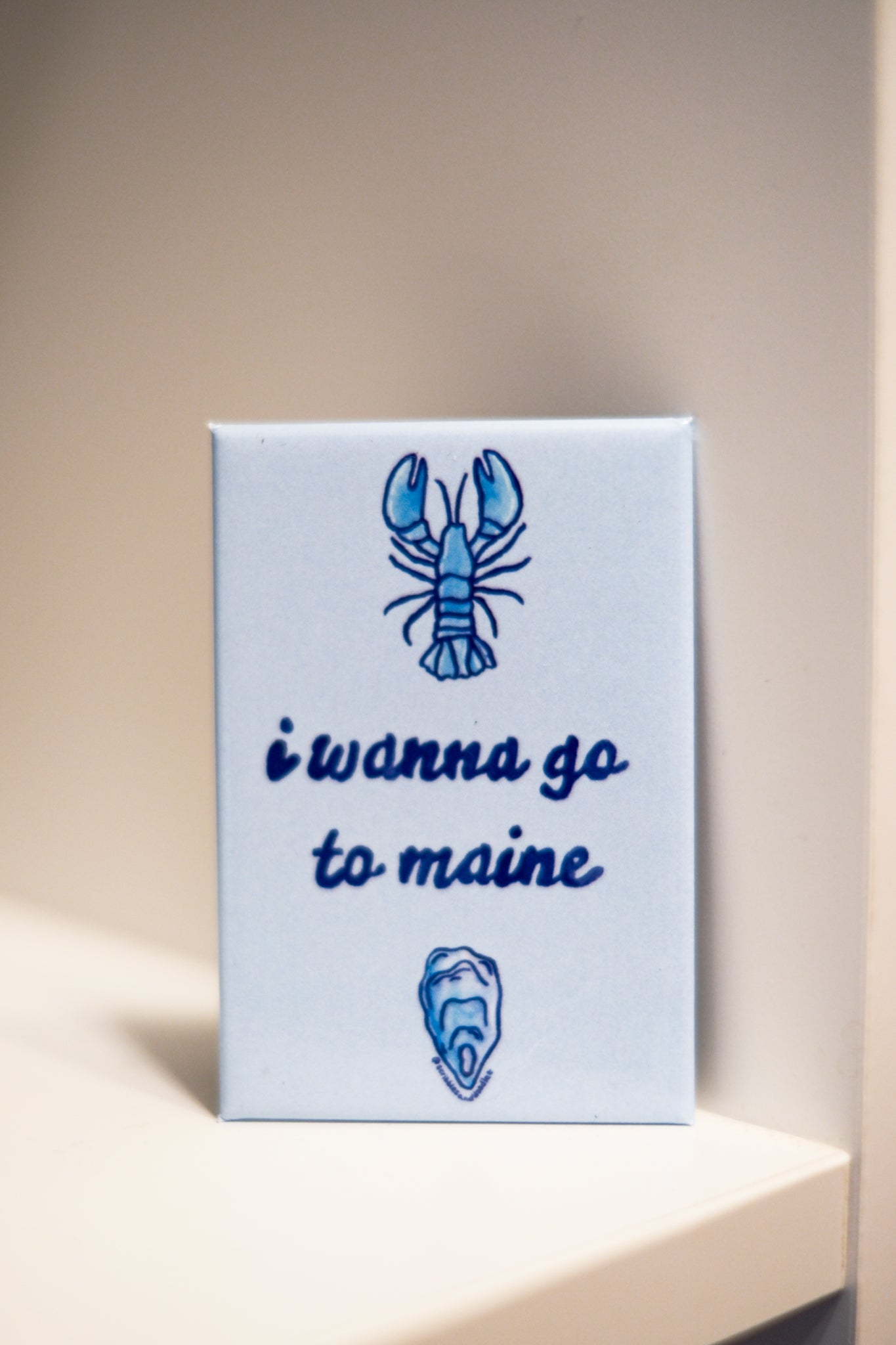 A square magnet printed with a blue lobster illustration 'i wanna go to maine' in dark blue script, and a small blue shell illustration.
