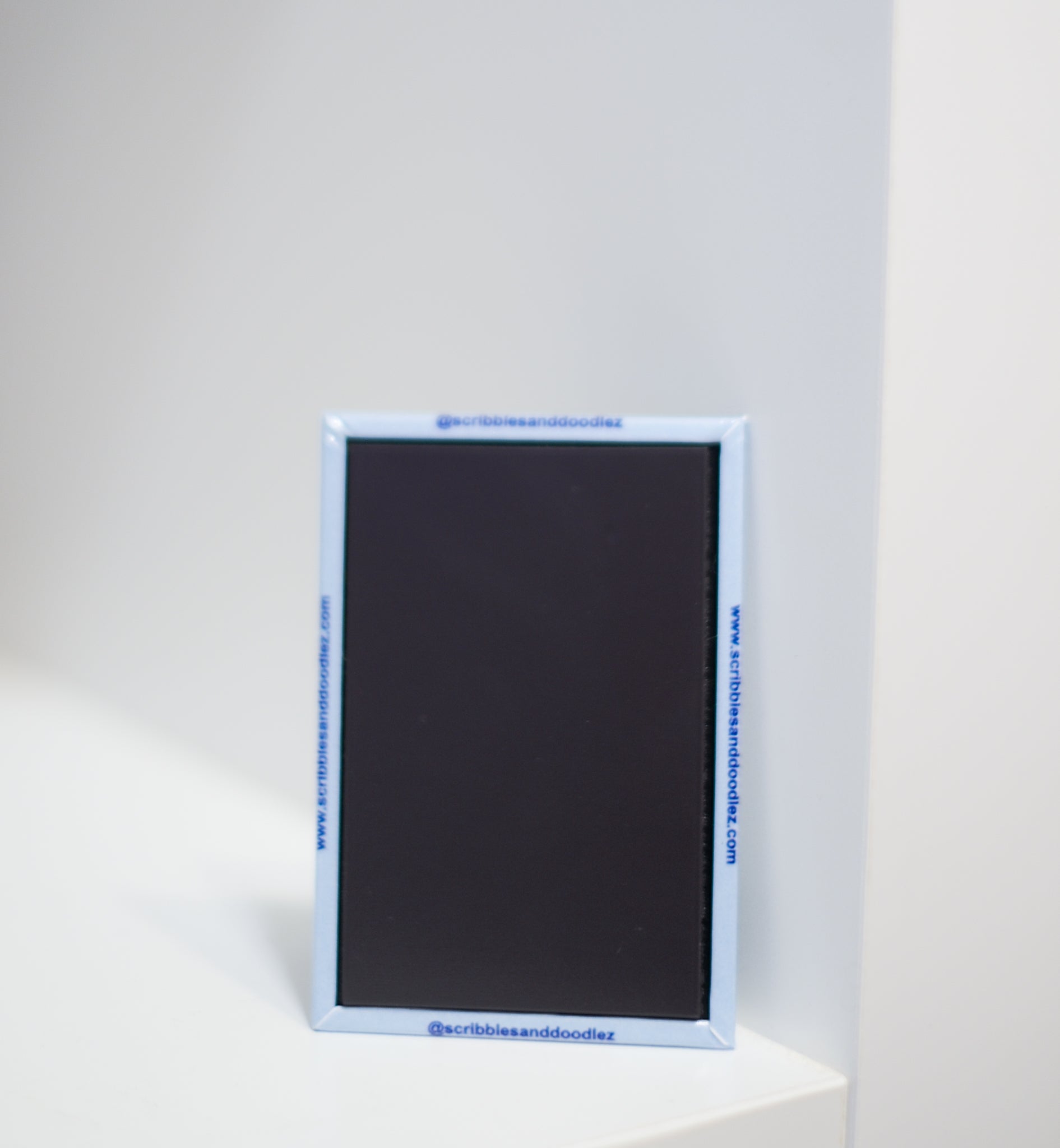 A small rectangular magnet with a light blue border stands upright on a white surface against a white background. Blue text is printed along the border, including 'www.scribblesanddoodlez.com' and '@scribblesanddoodlez'