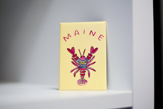 A yellow fridge magnet featuring a colorful lobster illustration and the word 'MAINE' in pink, standing upright on a white shelf against a gray background.