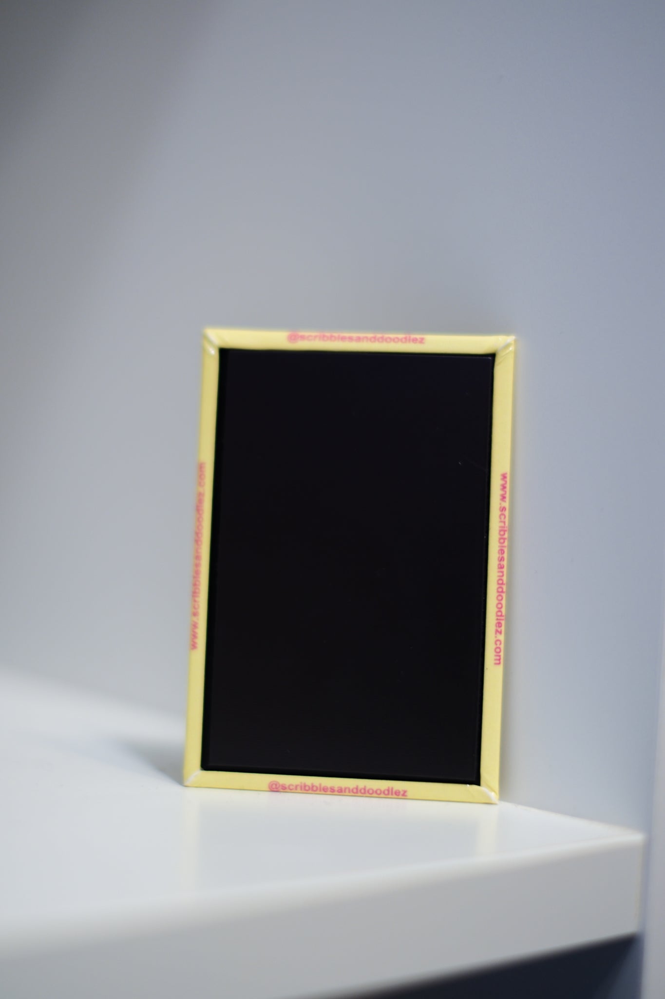 A small rectangular magnet with a yellow border, standing upright on a white surface against a light gray background, with the text in red '@scribblesanddoodlez' and 'www.scribblesanddoodlez.com'