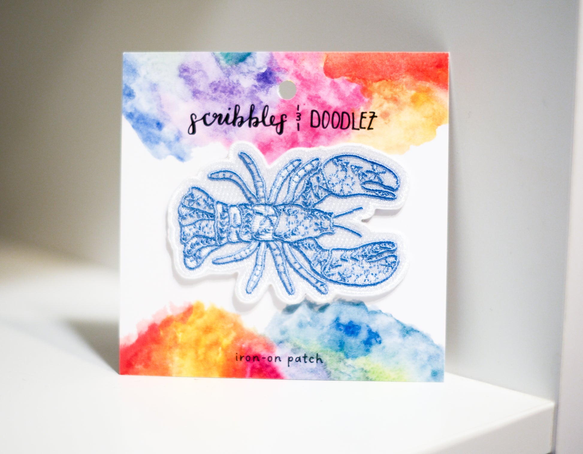 A white lobster shaped iron on patch with blue floral pattern displayed on its backing card, which features a colorful watercolor background and the brand name 'scribbles & doodlez.