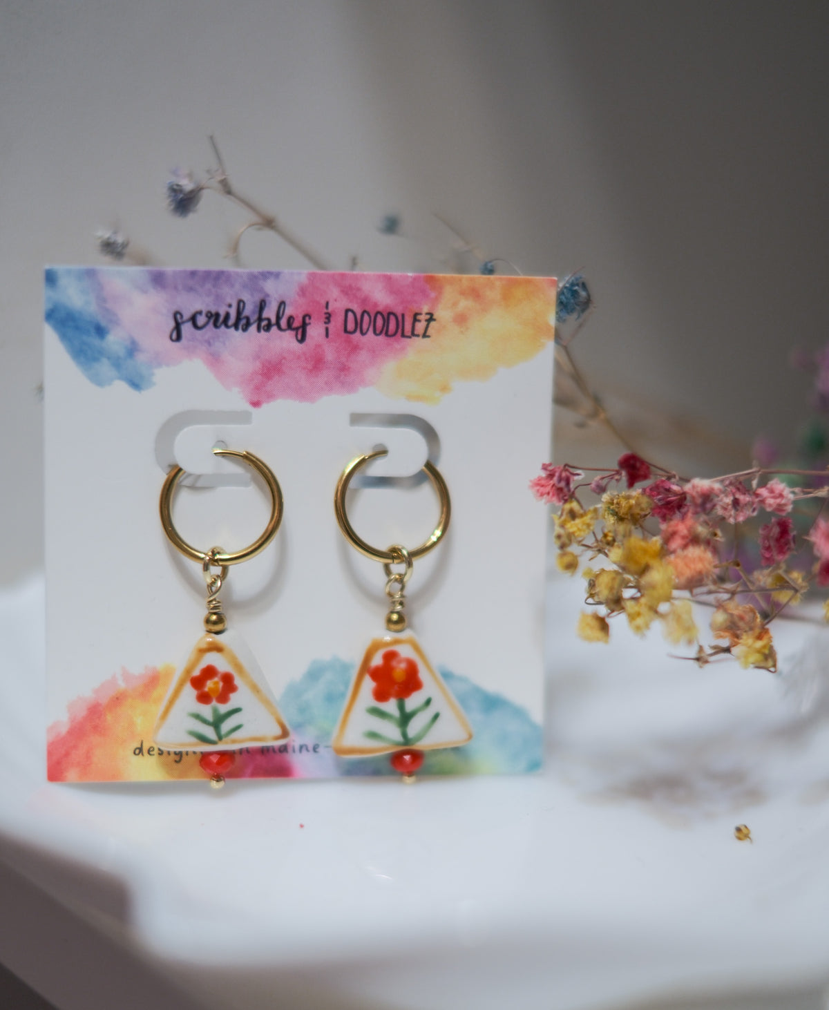 A pair of gold hoop earrings with triangular glazed ceramic bead featuring orange flowers and green stems, displayed on a colorful card labeled 'scribbles & doodlez' with dried flowers in the background.