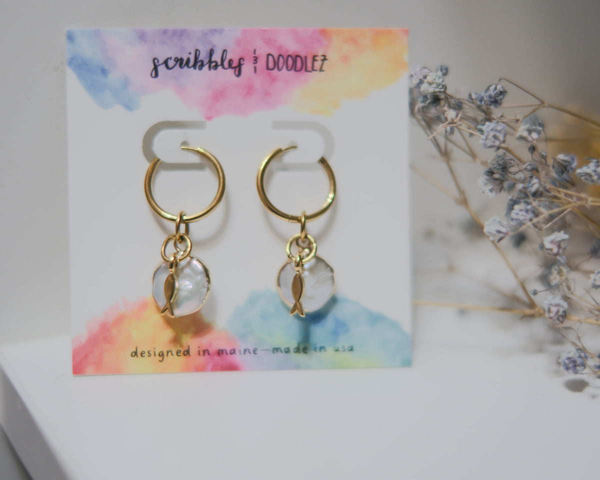 A pair of gold hoop earrings with pearl and tiny gold sardine charms, displayed on a white card with a colorful watercolor background. The card is branded 'scribbles + doodlez'.