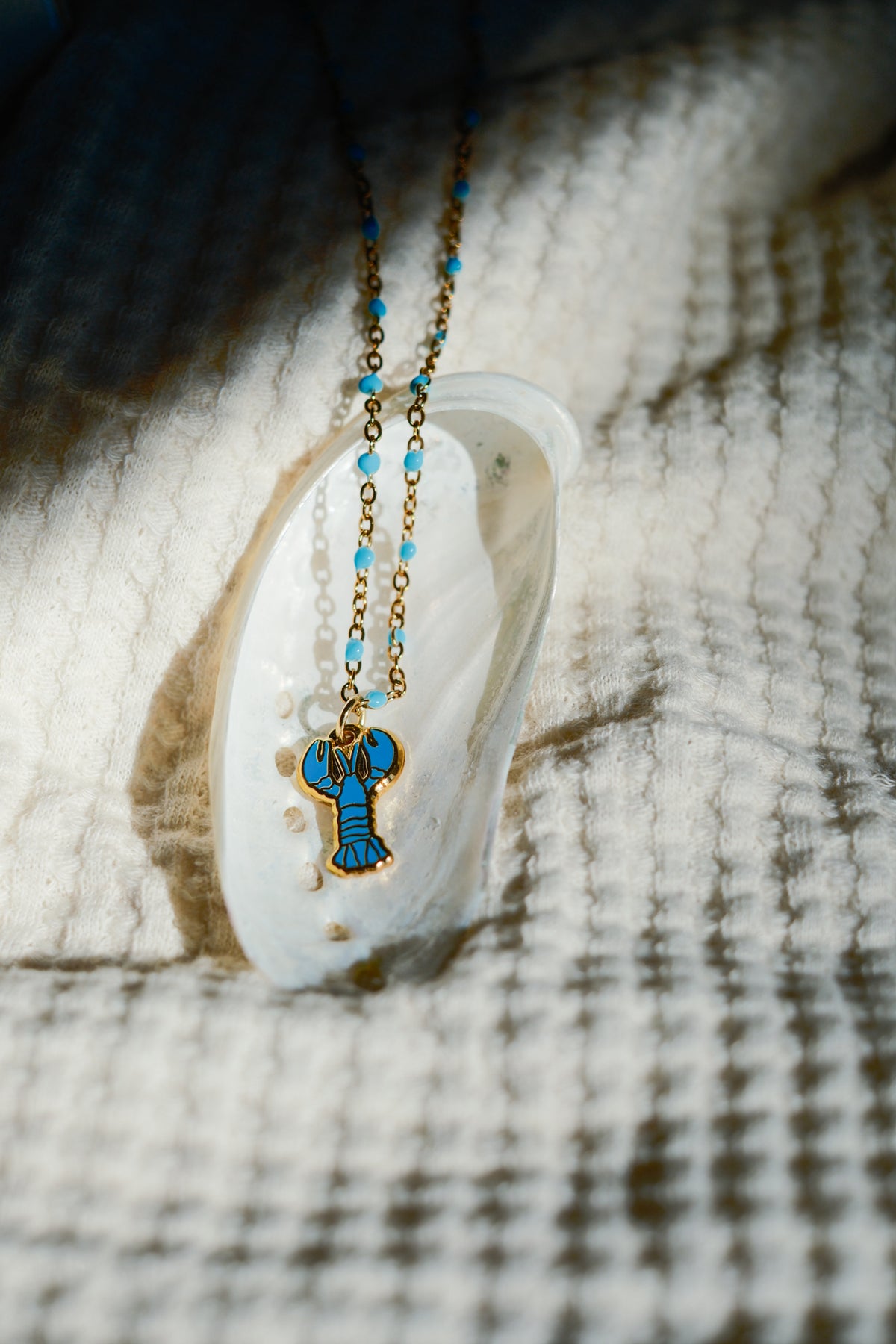 A gold-tone necklace with a blue enamel lobster pendant and small blue beads, displayed on a white clam shell over a textured beige woven blanket.