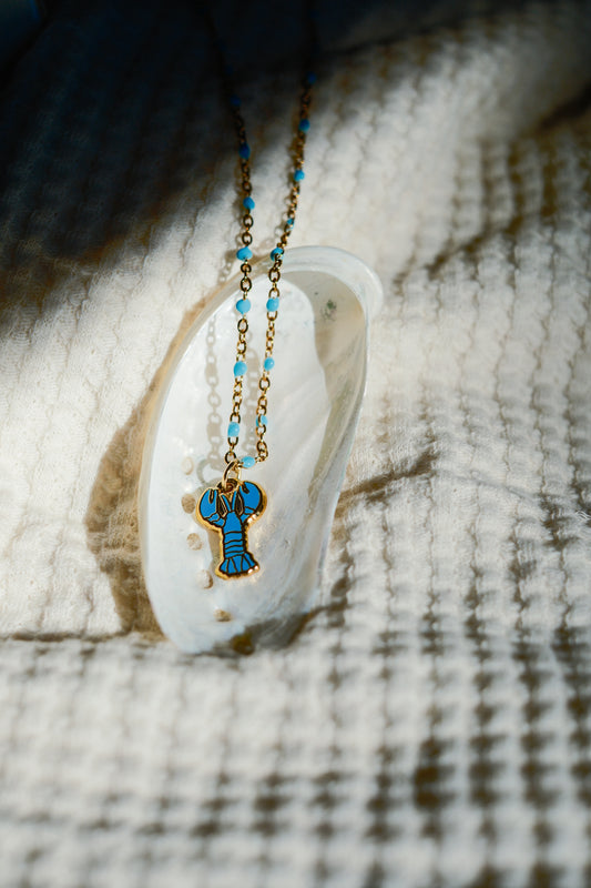 A gold-tone necklace with a blue enamel lobster pendant and small blue beads, displayed on a white clam shell over a textured beige woven blanket.